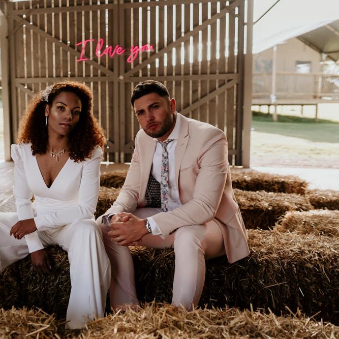 Black Bride with Afro Hair in Jumpsuit and Groom in Pink Suit Sitting On Hay Bales in a Barn
