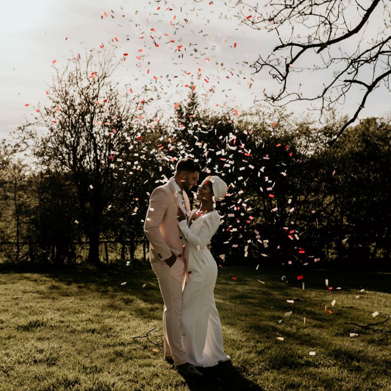 Confetti Moment with Black Bride in White Jumpsuit with Head Wrap  and Groom in Pink Suit