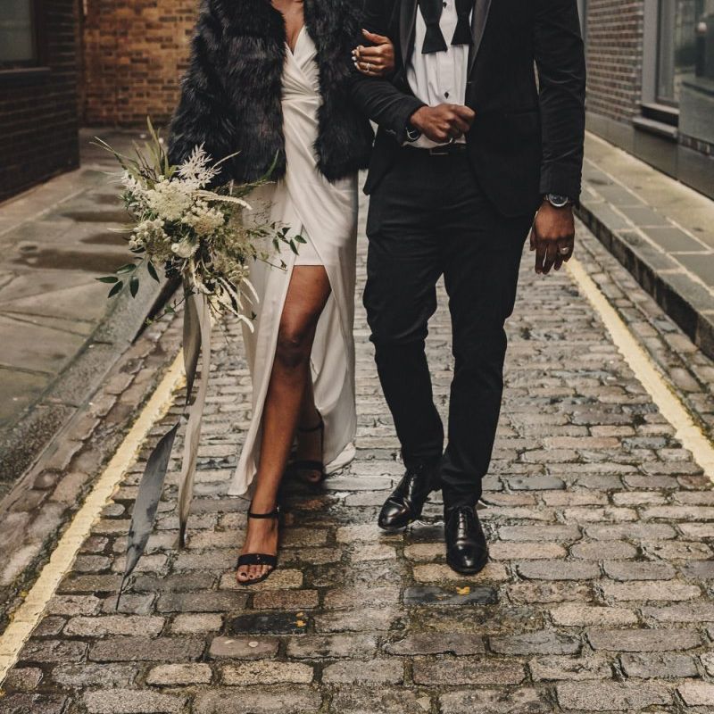 Stylish black bride in satin wedding dress and faux fur cover up and groom in bow tie and hat