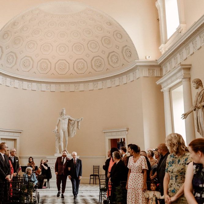 Groom in burgundy suit walking down the aisle with his father