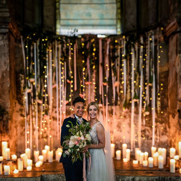 Portrait of Bride in Navy Suit and Pink Tie and Bride in Romantic Flora Mila Wedding Dress  Standing at The Asylum Altar Decorated in Candles and Ribbons
