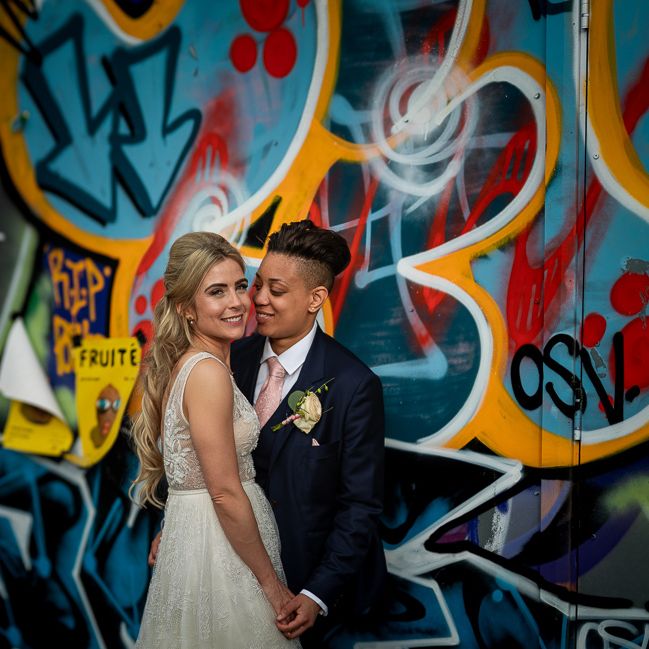Bride in Navy Suit and Pink Tie and Bride in Flora Mila Wedding Dress Embracing in Front of a Wall Muriel