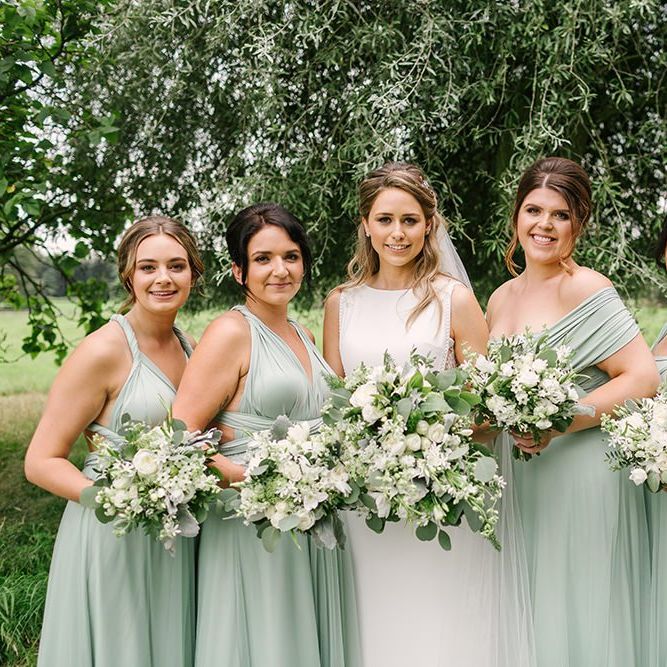 Bridal Party Portrait with Bridesmaids in Pale Green Multiway Dresses and Bride in Pronovias Wedding Dress Holding White Flower and Foliage Bouquets