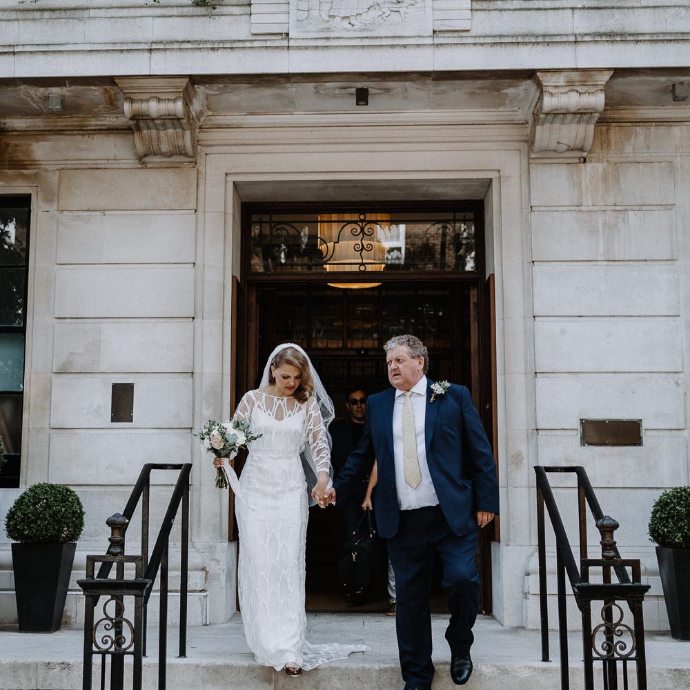 Bride in Embroidered Tulle Halfpenny London Dress with High Neckline, Batwing Sleeves and Low V Back | Fingertip Length Veil from Florence &amp; Dot | Bridal Bouquet of White and Blush Pink Flowers with Eucalyptus and Trailing Silk Ribbon | Father of the Bride in Navy Suit with Ivory Tie | Floral Arch and Bride in Batwing Sleeve Halfpenny Dress for London Pub Wedding | Caitlin + Jones