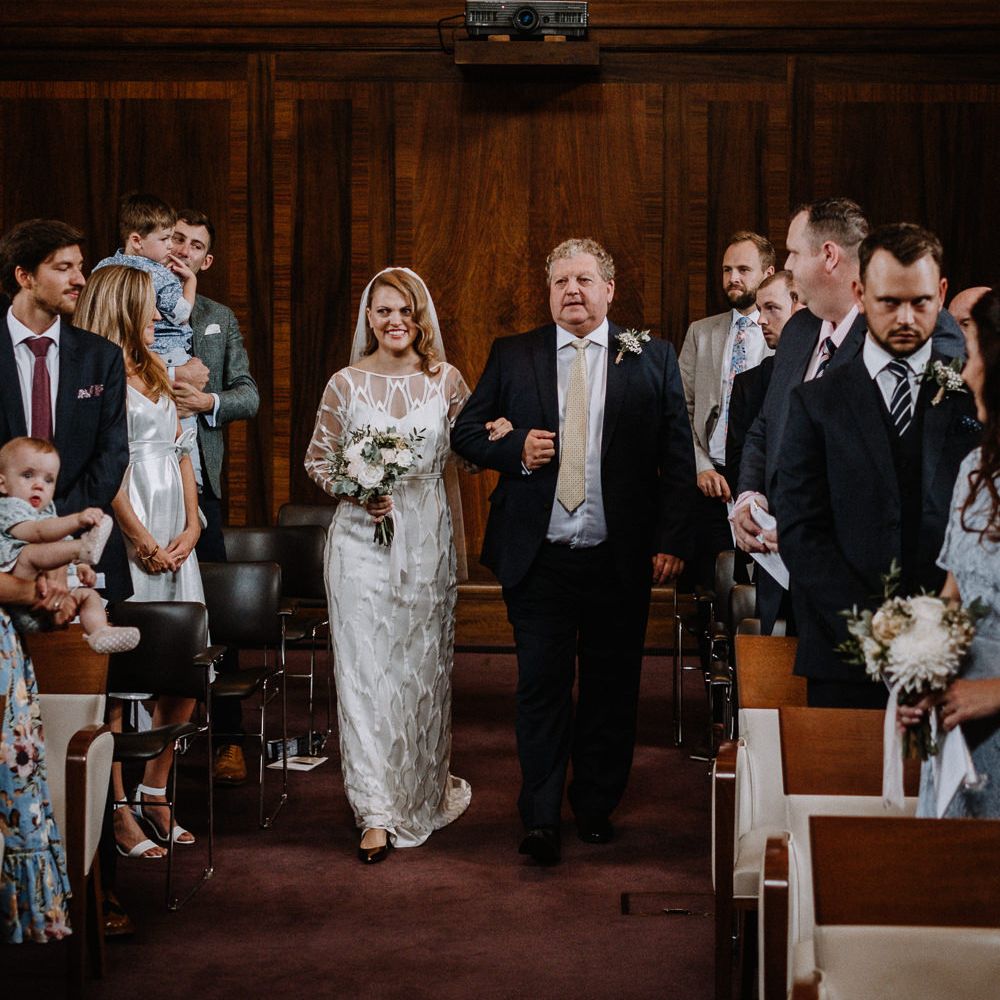 Bride in Embroidered Tulle Halfpenny London Dress with High Neckline, Batwing Sleeves and Low V Back | Fingertip Length Veil from Florence &amp; Dot | Bridal Bouquet of White and Blush Pink Flowers with Eucalyptus and Trailing Silk Ribbon | Father of the Bride in Navy Suit with Ivory Tie | Wedding Ceremony at Stoke Newington Town Hall | Floral Arch and Bride in Batwing Sleeve Halfpenny Dress for London Pub Wedding | Caitlin + Jones