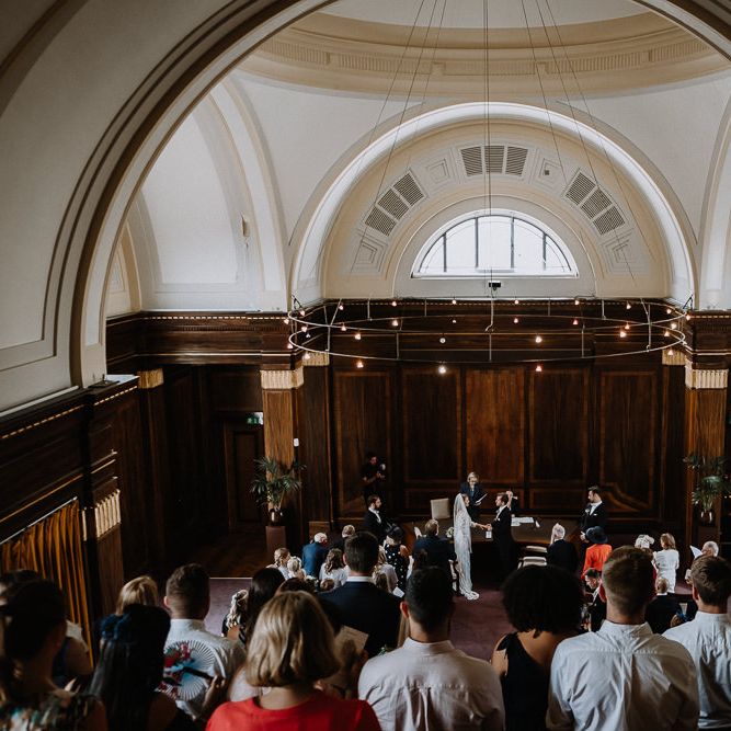 Bride in Embroidered Tulle Halfpenny London Dress with High Neckline, Batwing Sleeves and Low V Back | Fingertip Length Veil from Florence &amp; Dot | Groom in Navy Slim Fit Suit with Checked Double Breasted Waistcoat and Blush Pink Knitted Tie | Wedding Ceremony at Stoke Newington Town Hall | Floral Arch and Bride in Batwing Sleeve Halfpenny Dress for London Pub Wedding | Caitlin + Jones
