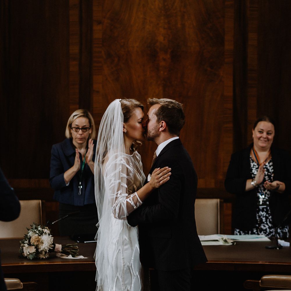 Bride in Embroidered Tulle Halfpenny London Dress with High Neckline, Batwing Sleeves and Low V Back | Fingertip Length Veil from Florence &amp; Dot | Bridal Bouquet of White and Blush Pink Flowers with Eucalyptus and Trailing Silk Ribbon | Groom in Navy Slim Fit Suit with Checked Double Breasted Waistcoat and Blush Pink Knitted Tie | Wedding Ceremony at Stoke Newington Town Hall | Floral Arch and Bride in Batwing Sleeve Halfpenny Dress for London Pub Wedding | Caitlin + Jones
