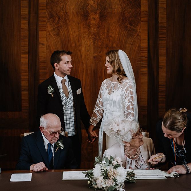 Bride in Embroidered Tulle Halfpenny London Dress with High Neckline, Batwing Sleeves and Low V Back | Fingertip Length Veil from Florence &amp; Dot | Bridal Bouquet of White and Blush Pink Flowers with Eucalyptus and Trailing Silk Ribbon | Groom in Navy Slim Fit Suit with Checked Double Breasted Waistcoat and Blush Pink Knitted Tie | Wedding Ceremony at Stoke Newington Town Hall | Floral Arch and Bride in Batwing Sleeve Halfpenny Dress for London Pub Wedding | Caitlin + Jones