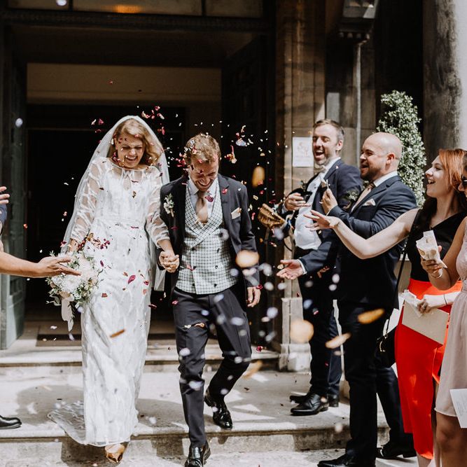 Bride in Embroidered Tulle Halfpenny London Dress with High Neckline, Batwing Sleeves and Low V Back | Fingertip Length Veil from Florence &amp; Dot | Bridal Bouquet of White and Blush Pink Flowers with Eucalyptus and Trailing Silk Ribbon | Groom in Navy Slim Fit Suit with Checked Double Breasted Waistcoat and Blush Pink Knitted Tie | Confetti Moment at Stoke Newington Town Hall | Floral Arch and Bride in Batwing Sleeve Halfpenny Dress for London Pub Wedding | Caitlin + Jones