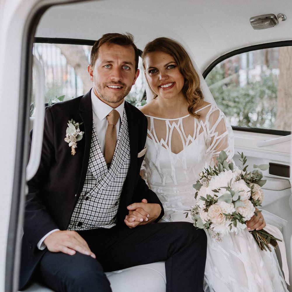Bride in Embroidered Tulle Halfpenny London Dress with High Neckline, Batwing Sleeves and Low V Back | Fingertip Length Veil from Florence &amp; Dot | Bridal Bouquet of White and Blush Pink Flowers with Eucalyptus and Trailing Silk Ribbon | Groom in Navy Slim Fit Suit with Checked Double Breasted Waistcoat and Blush Pink Knitted Tie | Wedding Ceremony at Stoke Newington Town Hall | Floral Arch and Bride in Batwing Sleeve Halfpenny Dress for London Pub Wedding | Caitlin + Jones