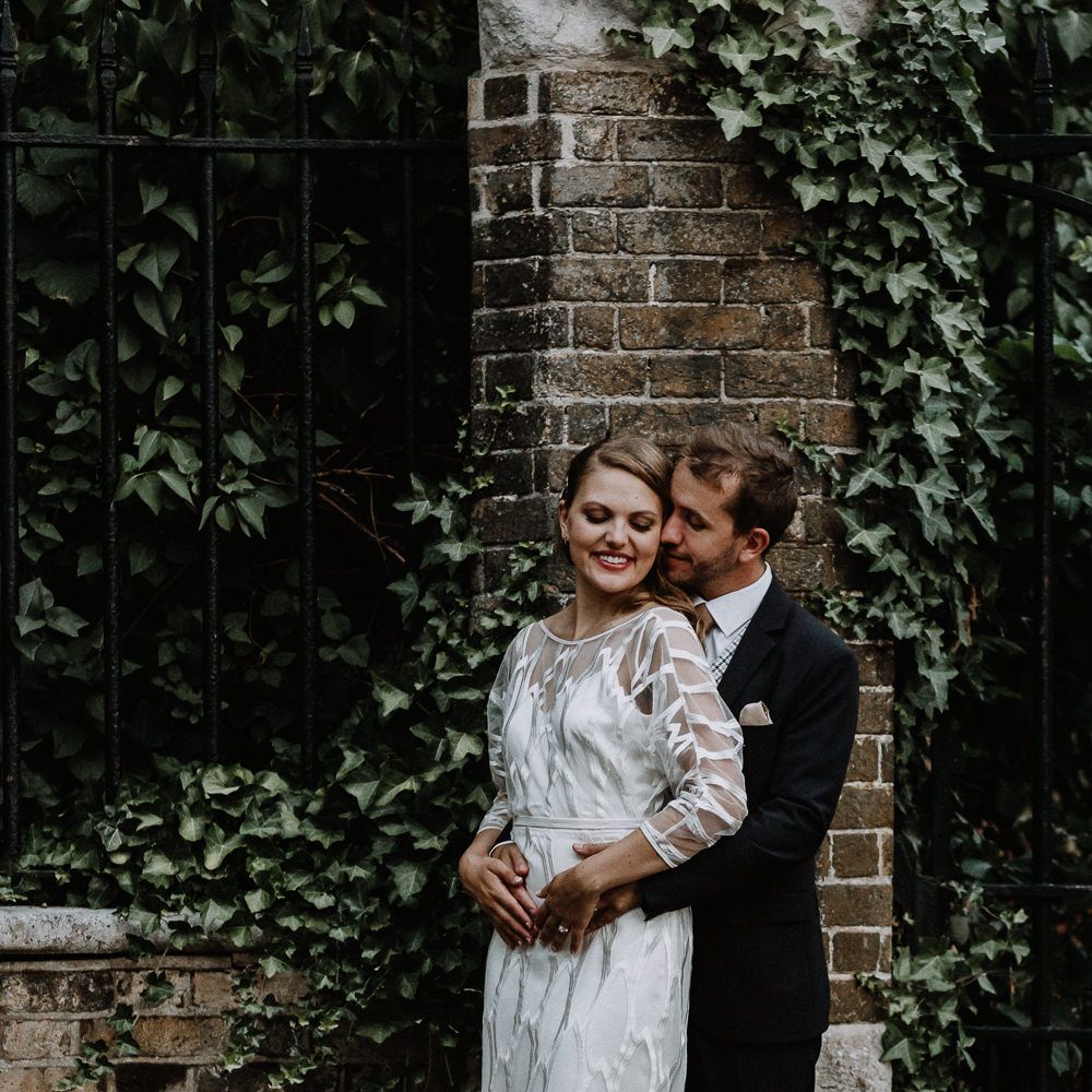 Bride in Embroidered Tulle Halfpenny London Dress with High Neckline, Batwing Sleeves and Low V Back | Groom in Navy Slim Fit Suit with Checked Double Breasted Waistcoat and Blush Pink Knitted Tie | Floral Arch and Bride in Batwing Sleeve Halfpenny Dress for London Pub Wedding | Caitlin + Jones