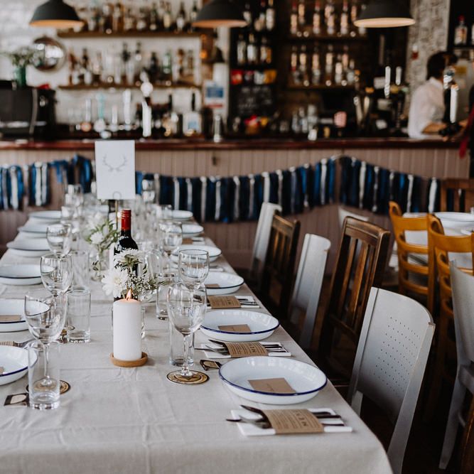 Banquet Tables | Mismatched Chairs | White Enamel Plates with Blue Rim | White Pillar Candle | Kraft Paper Wedding Breakfast Menus | Bud Vases with White Flowers and Eucalyptus | Blue and White Tassels | Floral Arch and Bride in Batwing Sleeve Halfpenny Dress for London Pub Wedding | Caitlin + Jones
