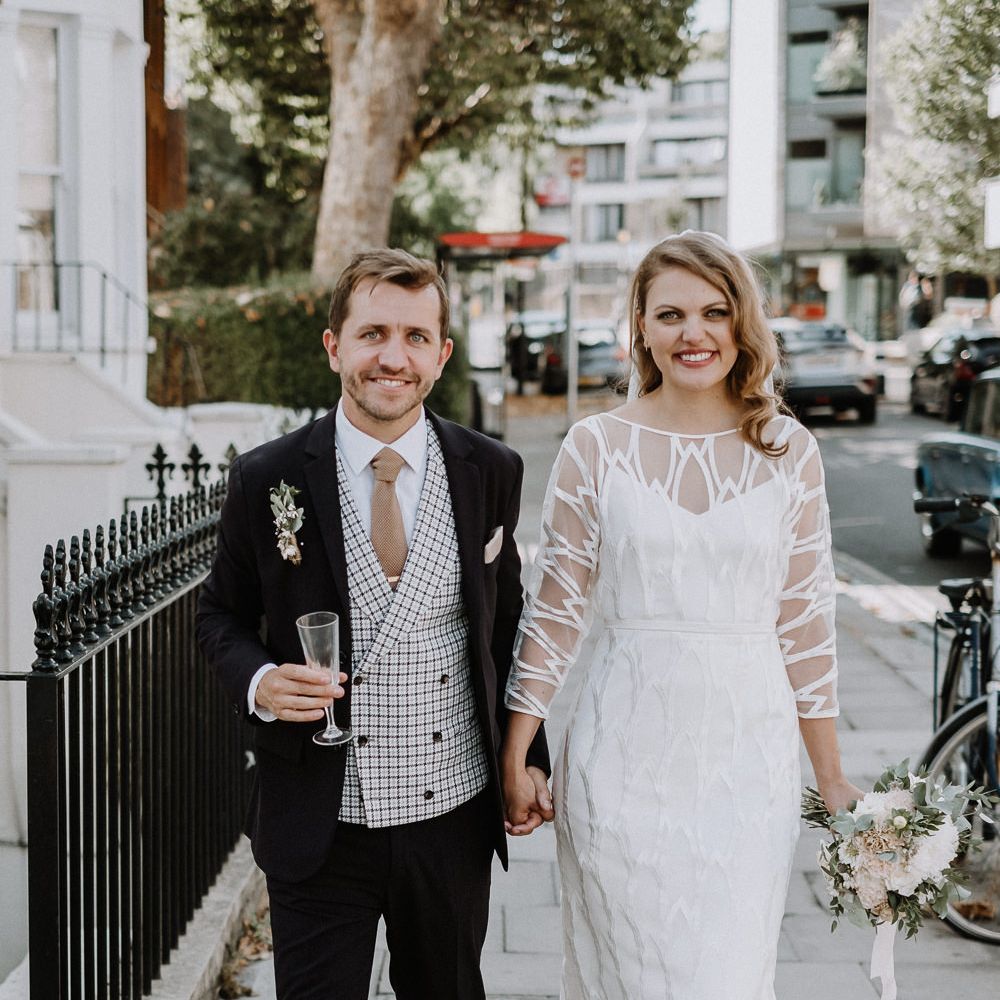 Bride in Embroidered Tulle Halfpenny London Dress with High Neckline, Batwing Sleeves and Low V Back | Fingertip Length Veil from Florence &amp; Dot | Bridal Bouquet of White and Blush Pink Flowers with Eucalyptus and Trailing Silk Ribbon | Groom in Navy Slim Fit Suit with Checked Double Breasted Waistcoat and Blush Pink Knitted Tie | Floral Arch and Bride in Batwing Sleeve Halfpenny Dress for London Pub Wedding | Caitlin + Jones