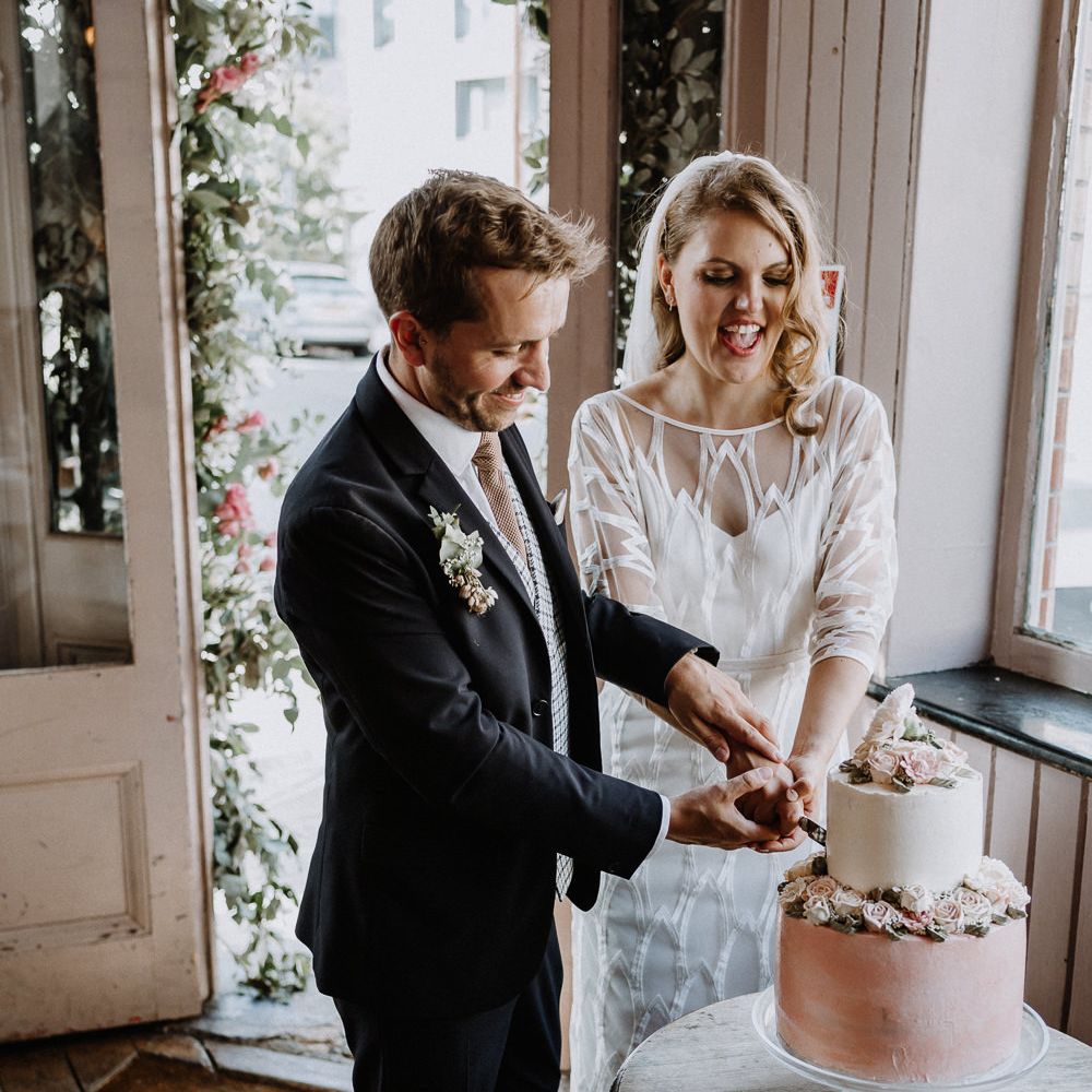 Two Tier Wedding Cake with Buttercream Icing and Iced Flowers | Bride in Embroidered Tulle Halfpenny London Dress with High Neckline, Batwing Sleeves and Low V Back | Fingertip Length Veil from Florence &amp; Dot | Groom in Navy Slim Fit Suit with Checked Double Breasted Waistcoat and Blush Pink Knitted Tie | Wedding Reception at The Star, Highgate | Floral Arch and Bride in Batwing Sleeve Halfpenny Dress for London Pub Wedding | Caitlin + Jones