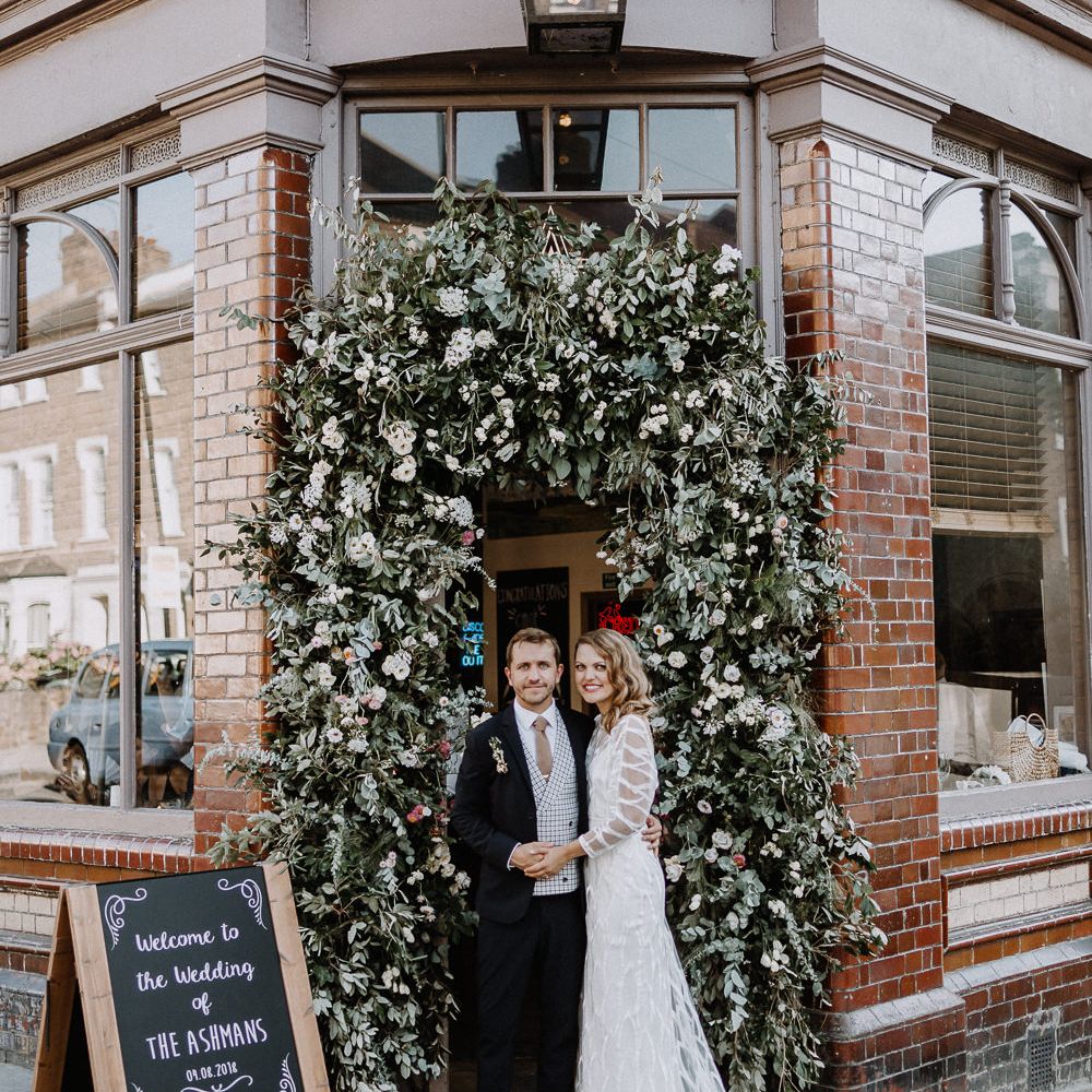 Bride in Embroidered Tulle Halfpenny London Dress with High Neckline, Batwing Sleeves and Low V Back | Groom in Navy Slim Fit Suit with Checked Double Breasted Waistcoat and Blush Pink Knitted Tie | Floral Arch | Chalkboard Welcome Sign | Wedding Reception at The Star Pub, Highgate | Floral Arch and Bride in Batwing Sleeve Halfpenny Dress for London Pub Wedding | Caitlin + Jones