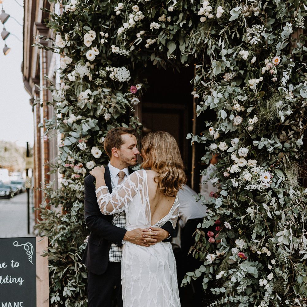 Bride in Embroidered Tulle Halfpenny London Dress with High Neckline, Batwing Sleeves and Low V Back | Groom in Navy Slim Fit Suit with Checked Double Breasted Waistcoat and Blush Pink Knitted Tie | Floral Arch | Chalkboard Welcome Sign | Wedding Reception at The Star Pub, Highgate | Floral Arch and Bride in Batwing Sleeve Halfpenny Dress for London Pub Wedding | Caitlin + Jones