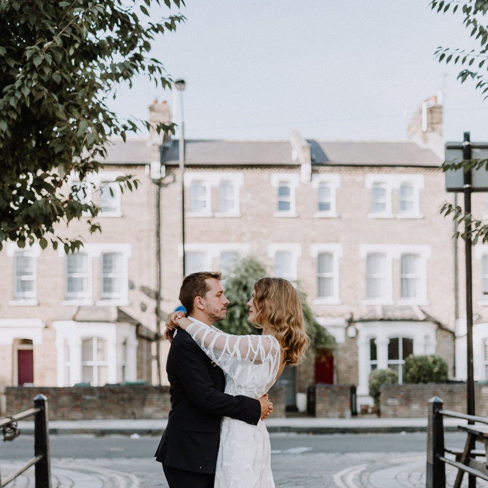 Bride in Embroidered Tulle Halfpenny London Dress with High Neckline, Batwing Sleeves and Low V Back | Groom in Navy Slim Fit Suit with Checked Double Breasted Waistcoat and Blush Pink Knitted Tie | Floral Arch and Bride in Batwing Sleeve Halfpenny Dress for London Pub Wedding | Caitlin + Jones