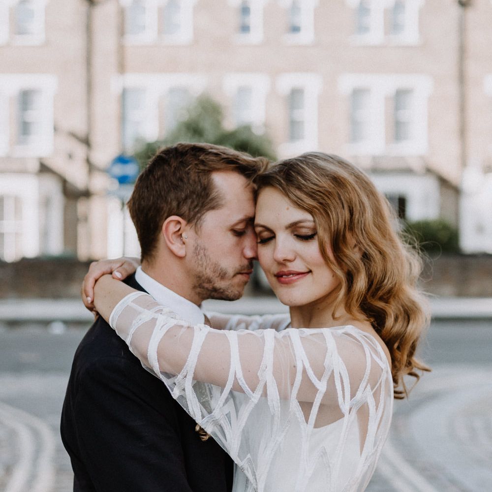 Bride in Embroidered Tulle Halfpenny London Dress with High Neckline, Batwing Sleeves and Low V Back | Groom in Navy Slim Fit Suit with Checked Double Breasted Waistcoat and Blush Pink Knitted Tie | Floral Arch and Bride in Batwing Sleeve Halfpenny Dress for London Pub Wedding | Caitlin + Jones