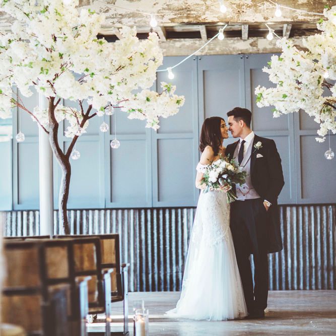 Bride in Fishtail Pronovias Wedding Dress with Off Shoulder Sleeves and Veil Cape | Groom in Navy Tails and Grey Waistcoat | Bride Wearing Hair Down | Bridal Bouquet of White Roses and Greenery | Cherry Blossom Tree Altar | Festoon Lights | Lace Bridal Cape Veil &amp; Fishtail Wedding Dress by Pronovias | On Love and Photography