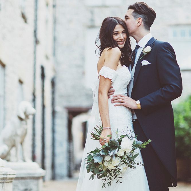 Bride in Fishtail Pronovias Wedding Dress with Off Shoulder Sleeves and Veil Cape | Groom in Navy Tails and Grey Waistcoat | Bride Wearing Hair Down | Bridal Bouquet of White Roses and Greenery | Lace Bridal Cape Veil &amp; Fishtail Wedding Dress by Pronovias | On Love and Photography