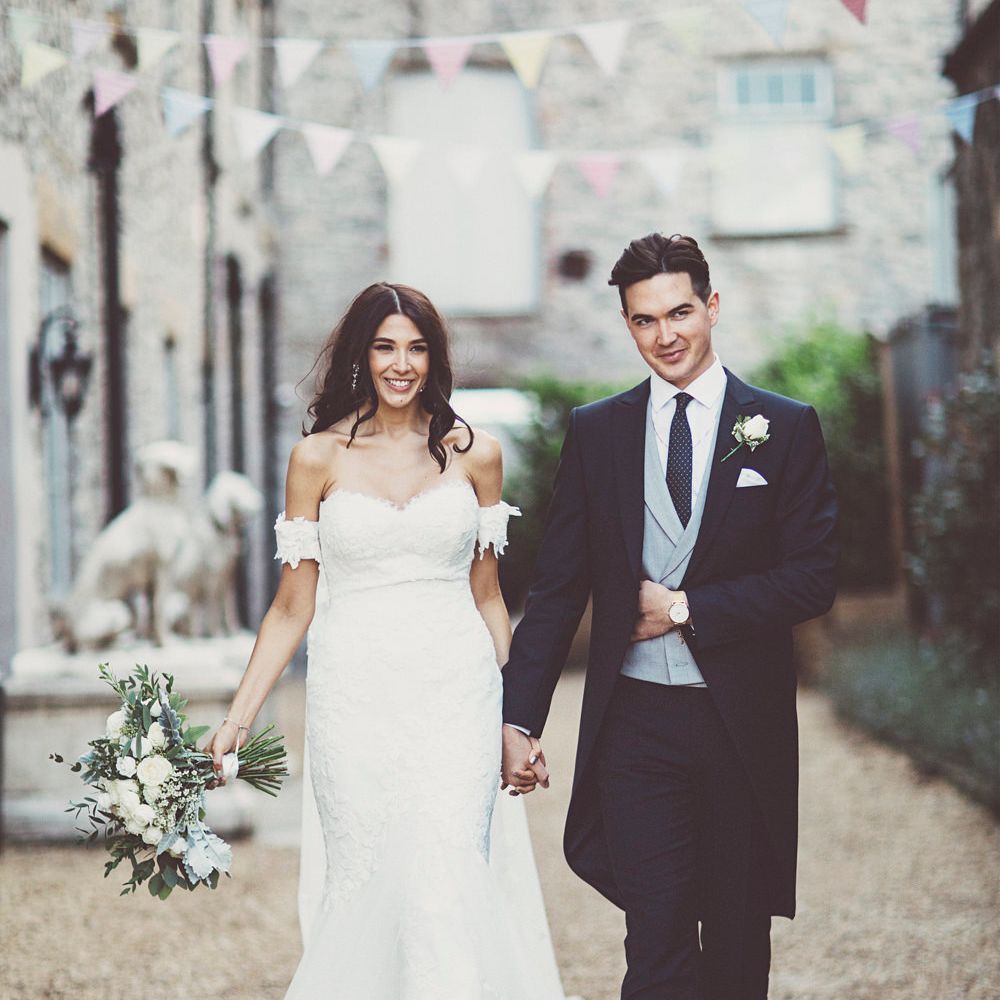 Bride in Fishtail Pronovias Wedding Dress with Off Shoulder Sleeves and Veil Cape | Groom in Navy Tails and Grey Waistcoat | Bride Wearing Hair Down | Bridal Bouquet of White Roses and Greenery | Lace Bridal Cape Veil &amp; Fishtail Wedding Dress by Pronovias | On Love and Photography