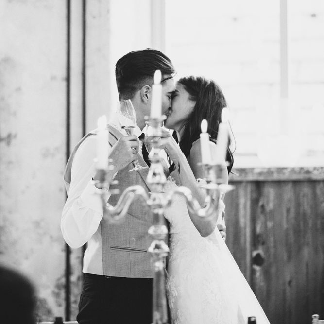 Newlywed Kiss | Bride in Fishtail Pronovias Wedding Dress with Off Shoulder Sleeves and Veil Cape | Groom in Navy Tails and Grey Waistcoat | Gold Candleabra with White Tapered Candles | Bride Wearing Hair Down | Exposed Copper Piping | Lace Bridal Cape Veil &amp; Fishtail Wedding Dress by Pronovias | On Love and Photography