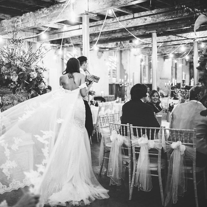 Bride in Fishtail Pronovias Wedding Dress with Off Shoulder Sleeves and Veil Cape | Groom in Navy Tails and Grey Waistcoat | Chalkboard Table Plan | White Material Chair Sashes | Oversized Floral Urn | Festoon Lights | Bride Wearing Hair Down | Lace Bridal Cape Veil &amp; Fishtail Wedding Dress by Pronovias | On Love and Photography