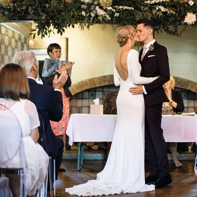 Bride in Long Sleeved Wedding Dress with Plunging V Back and Cuff Buttons by Pronovias | Groom in Black Three Piece Suit with Grey Tie and Personalised Tie Clip | Bridal Up Do | Wedding Altar Floral Installation | Wedding Ceremony Kiss | Ivania Pronovias Wedding Dress with Long Sleeves and Minimalist Styling | Chris Barber Photography