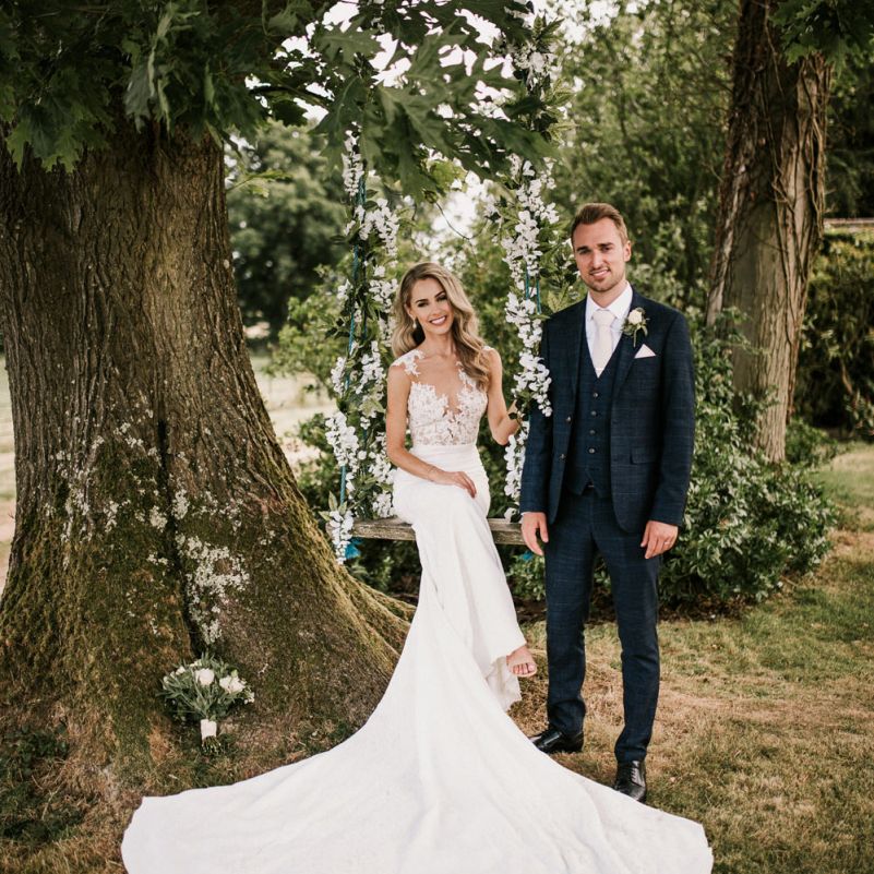 Bride in Pronovias Wedding Dress Sitting on a Ivy Covered Swing with Groom in Moss Bros. Suit  Standing By