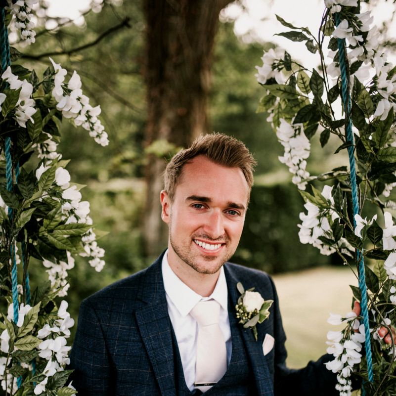 Groom in Navy Check Moss Bros. Suit Sitting on Ivy Covered Swing
