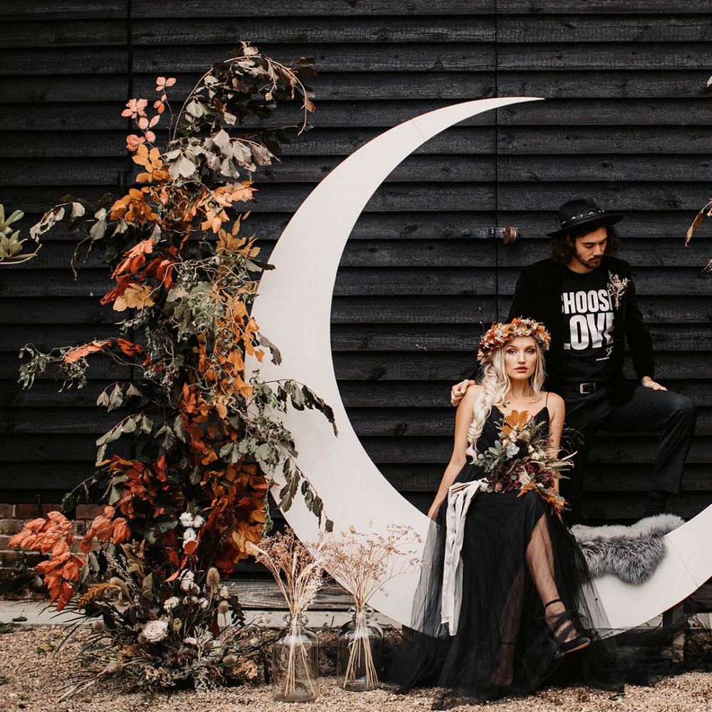 Stylish bride and groom sitting on a crescent moon flanked by autumnal floral arrangements  at Halloween wedding