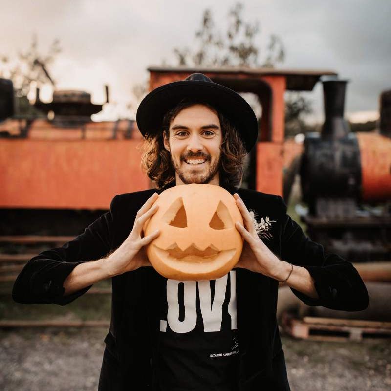 Groom in t-shirt, blazer and hat holding at carved pumpkin at Halloween wedding