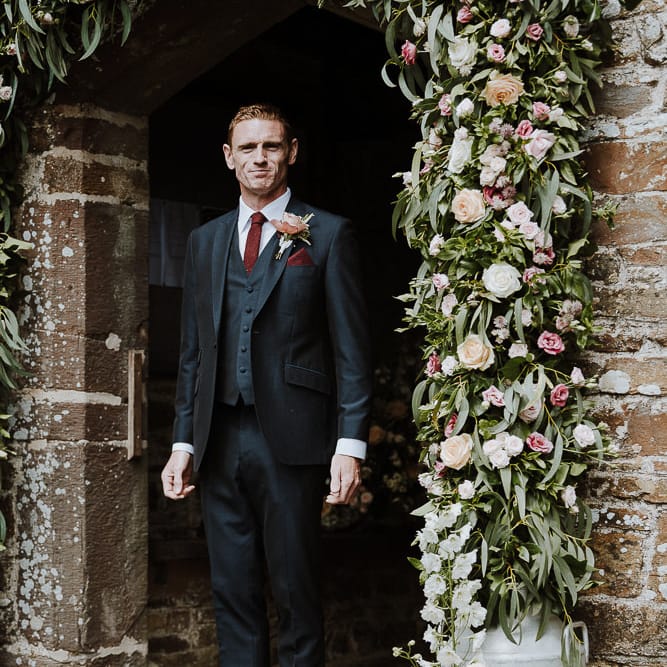 Groom in Three piece Clifton Suit Standing Next to a Church Floral Arch
