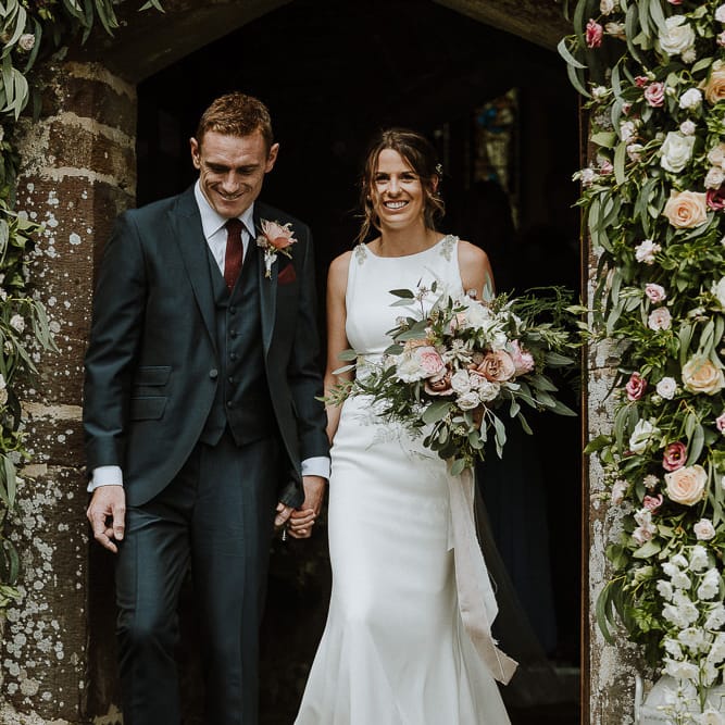 Bride in Mikaella Bridal Wedding Dress and Groom in Three piece Clifton Suit Exiting The Church Under a Floral Arch