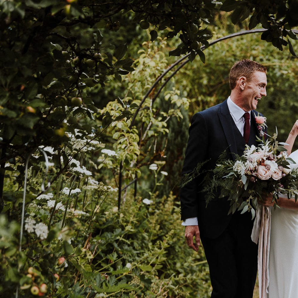 Bride in High Neck Mikaella Bridal Fitted Wedding Dress and Groom in  Clifton Suit Laughing
