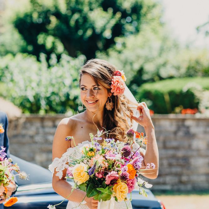 Bride in Structured Bustier Rue de Seine Fox Gown with Floral Appliqué Detail and Off The Shoulder Straps | Floor Length Veil | Bouquet of Bright Wild Flowers with White Ribbon | Rose Gold Earrings | Pink Dahlia Hair Flower | Father of the Bride in Blue Two Piece Suit with Tan Waistcoat and Bow Tie | Bridesmaid in White ASOS Dress | Colourful Bunting Wedding Decor for a Festival Themed Marquee Wedding | Peppermint Love Photography