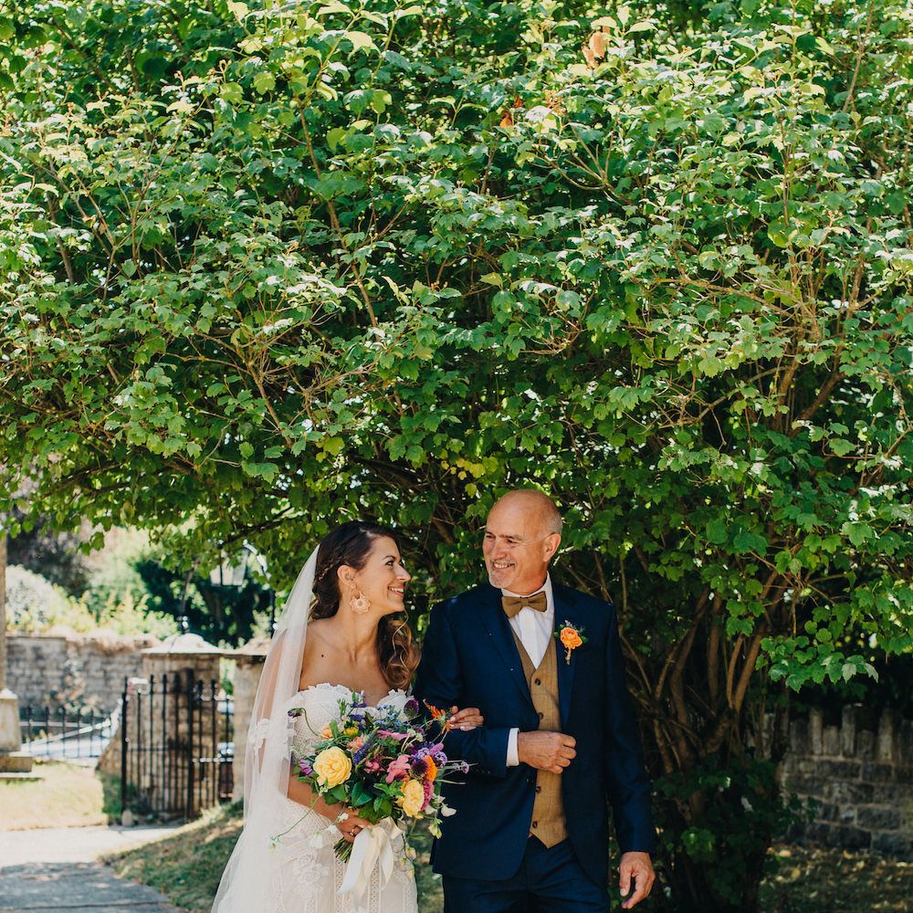 Bride in Structured Bustier Rue de Seine Fox Gown with Floral Appliqué Detail and Off The Shoulder Straps | Floor Length Veil | Rose Gold Earrings | Bouquet of Bright Wild Flowers with White Ribbon | Father of the Bride in Blue Two Piece Suit with Tan Waistcoat and Bow Tie | Colourful Bunting Wedding Decor for a Festival Themed Marquee Wedding | Peppermint Love Photography