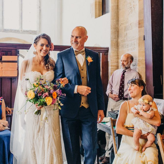 Bride in Structured Bustier Rue de Seine Fox Gown with Floral Appliqué Detail and Off The Shoulder Straps | Floor Length Veil | Rose Gold Earrings | Bouquet of Bright Wild Flowers with White Ribbon | Father of the Bride in Blue Two Piece Suit with Tan Waistcoat and Bow Tie | Entrance of the Bride | Colourful Bunting Wedding Decor for a Festival Themed Marquee Wedding | Peppermint Love Photography