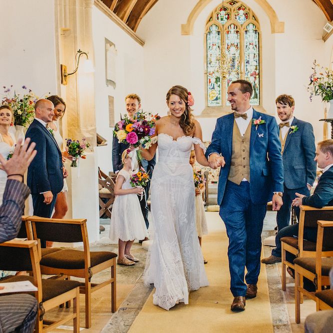 Bride in Structured Bustier Rue de Seine Fox Gown with Floral Appliqué Detail and Off The Shoulder Straps | Floor Length Veil | Rose Gold Earrings | Bouquet of Bright Wild Flowers with White Ribbon | Pink Dahlia Hair Flower | Groom in Blue Two Piece Suit with Tan Waistcoat and Bow Tie | Wedding Ceremony | Colourful Bunting Wedding Decor for a Festival Themed Marquee Wedding | Peppermint Love Photography