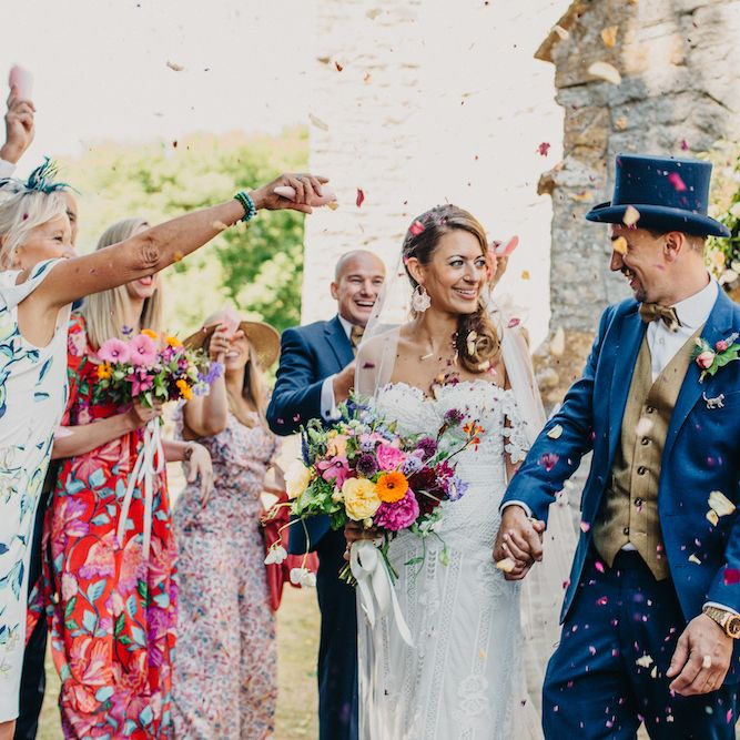 Bride in Structured Bustier Rue de Seine Fox Gown with Floral Appliqué Detail and Off The Shoulder Straps | Floor Length Veil | Rose Gold Earrings | Bouquet of Bright Wild Flowers with White Ribbon | Groom in Blue Two Piece Suit with Tan Waistcoat, Bow Tie and Top Hat | Confetti Moment | Colourful Bunting Wedding Decor for a Festival Themed Marquee Wedding | Peppermint Love Photography