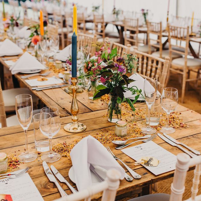 Wooden Banquet Tables | Colourful Tapered Candles in Gold Candlesticks | Scattered Dried Petals | Honey Wedding Favours | Colourful Bunting Wedding Decor for a Festival Themed Marquee Wedding | Peppermint Love Photography