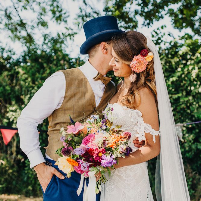 Bride in Structured Bustier Rue de Seine Fox Gown with Floral Appliqué Detail and Off The Shoulder Straps | Floor Length Veil | Rose Gold Earrings | Bouquet of Bright Wild Flowers with White Ribbon | Pink Dahlia Hair Flower | Groom in Blue Two Piece Suit with Tan Waistcoat, Bow Tie and Top Hat | Colourful Bunting Wedding Decor for a Festival Themed Marquee Wedding | Peppermint Love Photography