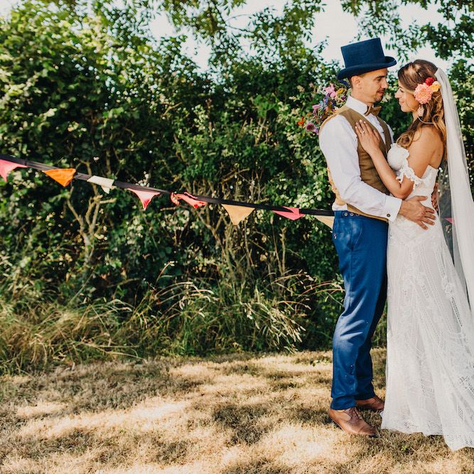Bride in Structured Bustier Rue de Seine Fox Gown with Floral Appliqué Detail and Off The Shoulder Straps | Floor Length Veil | Rose Gold Earrings | Pink Dahlia Hair Flower | Groom in Blue Two Piece Suit with Tan Waistcoat, Bow Tie and Top Hat | Colourful Bunting Wedding Decor for a Festival Themed Marquee Wedding | Peppermint Love Photography
