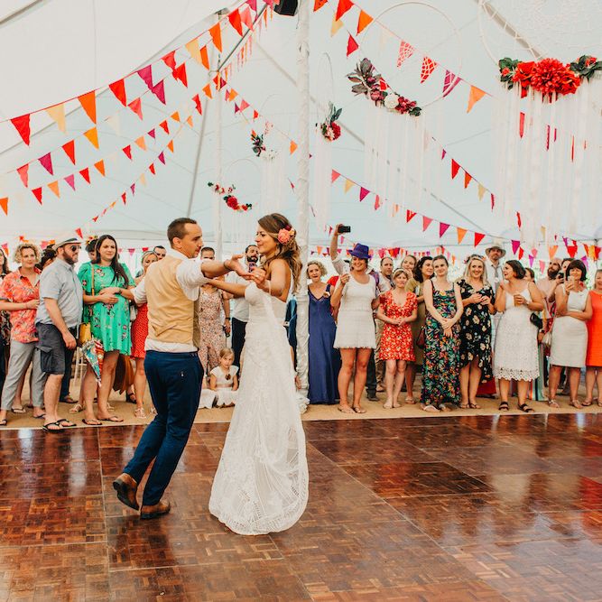 Bride in Structured Bustier Rue de Seine Fox Gown with Floral Appliqué Detail and Off The Shoulder Straps | Rose Gold Earrings | Pink Dahlia Hair Flowers | Groom in Blue Two Piece Suit with Tan Waistcoat and Bow Tie | First Dance | Colourful Bunting Wedding Decor for a Festival Themed Marquee Wedding | Peppermint Love Photography