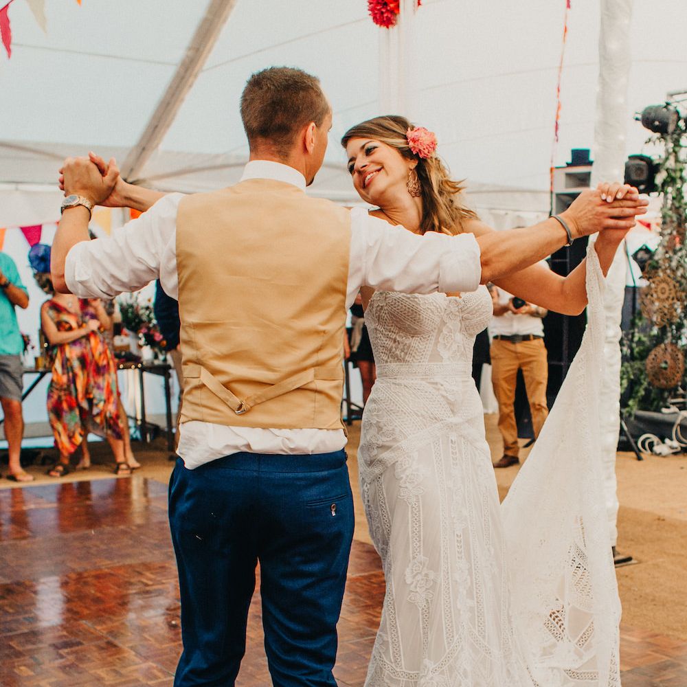 Bride in Structured Bustier Rue de Seine Fox Gown with Floral Appliqué Detail and Off The Shoulder Straps | Rose Gold Earrings | Pink Dahlia Hair Flowers | Groom in Blue Two Piece Suit with Tan Waistcoat and Bow Tie | First Dance | Colourful Bunting Wedding Decor for a Festival Themed Marquee Wedding | Peppermint Love Photography