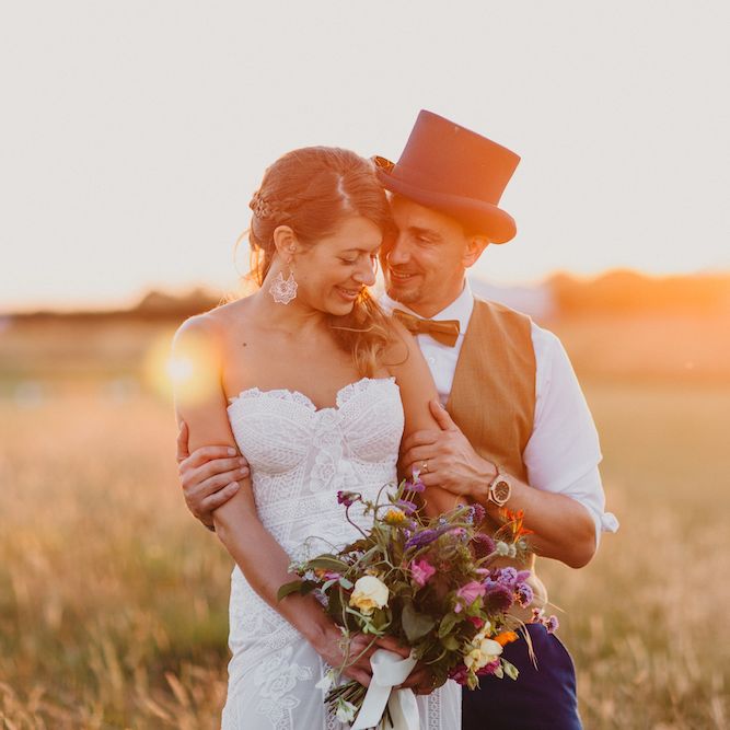 Bride in Structured Bustier Rue de Seine Fox Gown with Floral Appliqué Detail and Off The Shoulder Straps | Rose Gold Earrings | Bouquet of Bright Wild Flowers with White Ribbon | Pink Dahlia Hair Flower | Groom in Blue Two Piece Suit with Tan Waistcoat, Bow Tie and Top Hat | Colourful Bunting Wedding Decor for a Festival Themed Marquee Wedding | Peppermint Love Photography