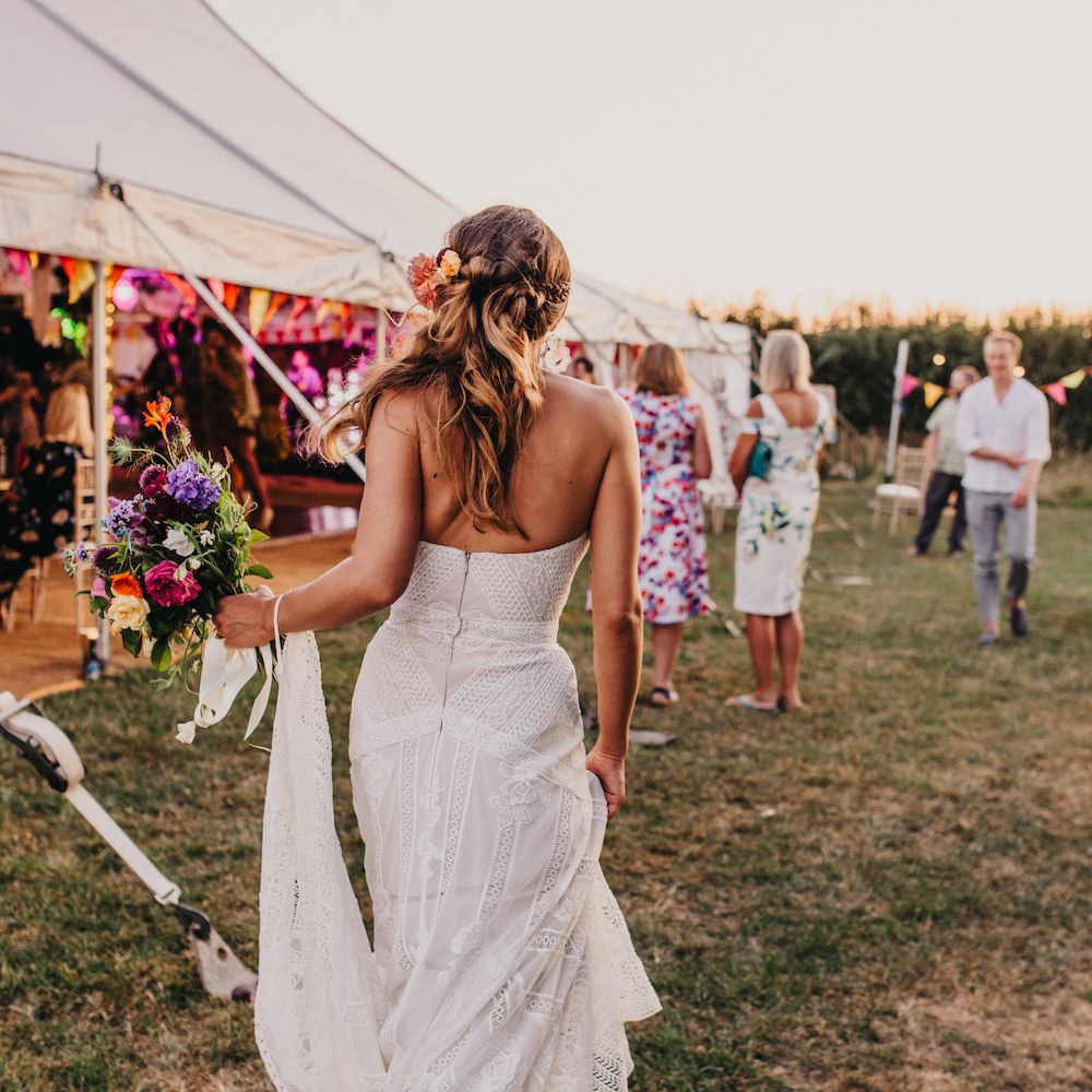 Bride in Structured Bustier Rue de Seine Fox Gown with Floral Appliqué Detail and Off The Shoulder Straps | Bouquet of Bright Wild Flowers with White Ribbon | Pink Dahlia Hair Flower | Colourful Bunting Wedding Decor for a Festival Themed Marquee Wedding | Peppermint Love Photography