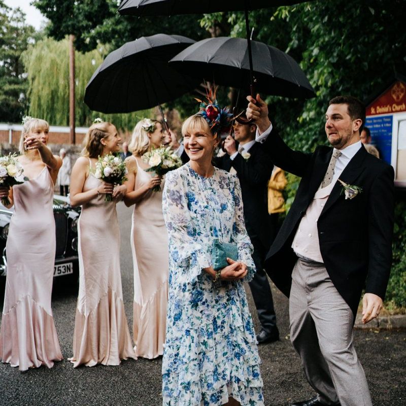 Mother of the bride and bridesmaids stand, smiling, underneath umbrellas as they wait for the bride to exit her car