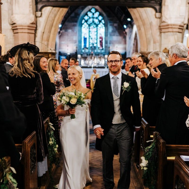 Bride and groom hold hands and walk back down the aisle as husband and wife while smiling at their guests ]