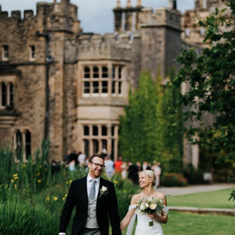 Bride and groom walk towards camera with Hawarden Castle in the background